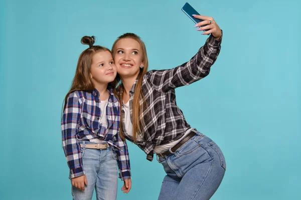 Mom and daughter dressed in checkered shirts and blue denim jeans are making selfie while posing against a blue studio background. Close-up shot.