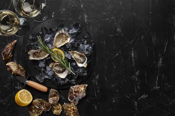 Las ostras frescas, el hielo, el limón en una pizarra redonda y el champán están en un fondo texturizado de piedra negra. Vista superior con espacio de copia. Cierre. —  Fotos de Stock