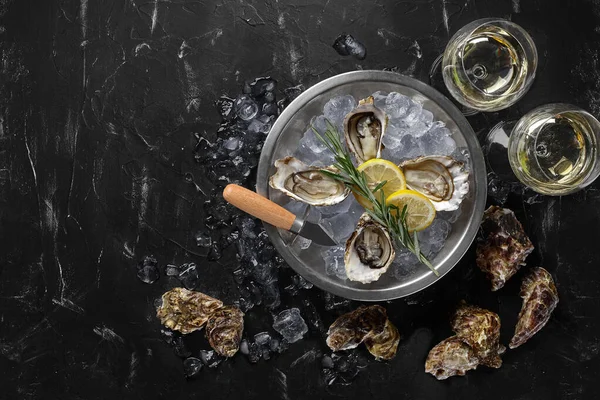 Fresh opened oysters, ice, lemon on a round metal plate and champagne are on a black stone textured background. Top view with copy space. Close-up. — Stock Photo, Image