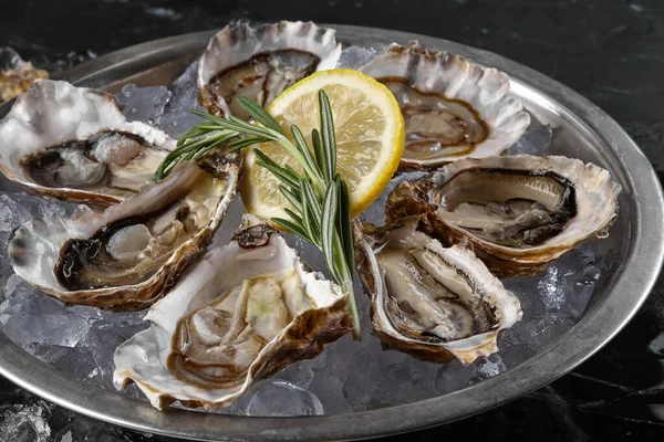 Fresh opened oysters, ice and lemon on a round metal plate, black stone textured background. Side view with copy space. Close-up shot. — Stock Photo, Image