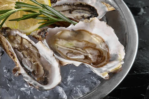 Fresh opened oysters, ice and lemon on a round metal plate, black stone textured background. Top view. Close-up shot. — Stock Photo, Image