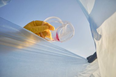 Sarı lastik eldivenli, bilinci açık birinin eli kullanılmış plastik bardağı mavi bir çöp torbasına koyar. Mavi gökyüzü. Nehir kenarı kirliliği. Yakın plan..