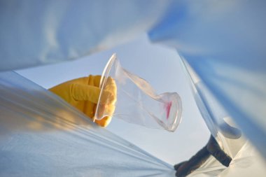 Sarı lastik eldivenli, bilinci açık birinin eli kullanılmış plastik bardağı mavi bir çöp torbasına koyar. Mavi gökyüzü. Nehir kenarı kirliliği. Yakın plan..