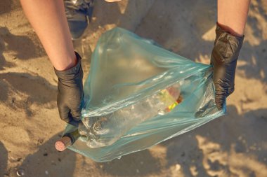 Siyah eldivenli genç gönüllü nehrin kirli sahilinde çöp torbasıyla yürüyor ve çöpleri temizliyor. İnsanlar ve ekoloji. Yakın plan..