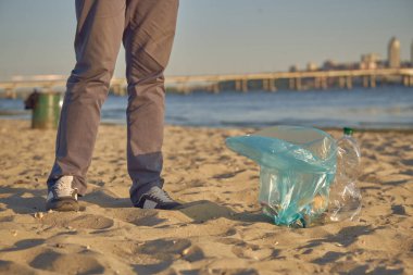 Genç gönüllü, nehrin kirli sahilinde çöp torbasıyla yürüyor ve çöpleri temizliyor. İnsanlar ve ekoloji. Yakın plan..