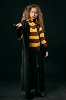 Siyah ceketli, uzun kahverengi saçlı, elinde sihirli değnekle siyah stüdyo arkasında poz veren küçük bir cadı kızın portresi..