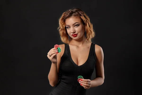 Ginger meisje het dragen van donkere jurk is poseren met rode en groene chips in haar handen staan tegen zwarte studio achtergrond. Casino, poker. Close-up. — Stockfoto