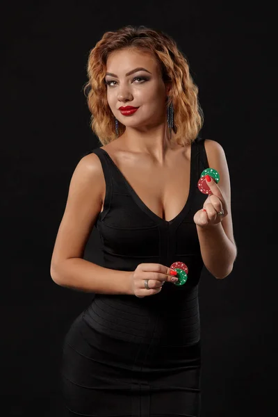 Ginger dívka v tmavých šatech je pózování drží červené a zelené čipy v rukou stojí na černém pozadí studia. Kasino, poker. Detailní záběr. — Stock fotografie
