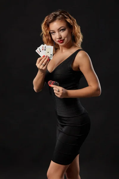 Ragazza zenzero in un abito scuro posa in possesso di carte da gioco e patatine fritte nelle sue mani in piedi su uno sfondo nero studio. Casino ', poker. Primo piano . — Foto Stock