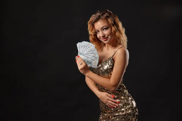 Ginger girl in shiny dress posing holding fan of hundred-dollar bills in her hands standing against black studio background. Casino, poker. Close-up.