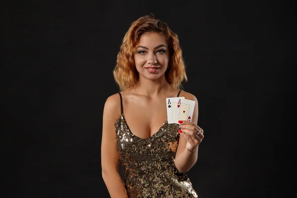 Ginger dívka na sobě lesklé šaty je pózování drží dvě hrací karty v rukou stojí na černém pozadí studia. Kasino, poker. Detailní záběr. — Stock fotografie