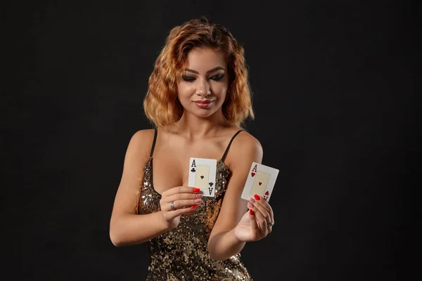 Ginger ragazza indossa abito lucido è in posa in possesso di due carte da gioco in mano in piedi su uno sfondo nero studio. Casino ', poker. Primo piano . — Foto Stock