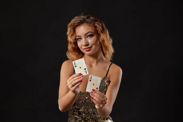 Ginger ragazza indossa abito lucido è in posa in possesso di due carte da gioco in mano in piedi su uno sfondo nero studio. Casino ', poker. Primo piano . — Foto Stock