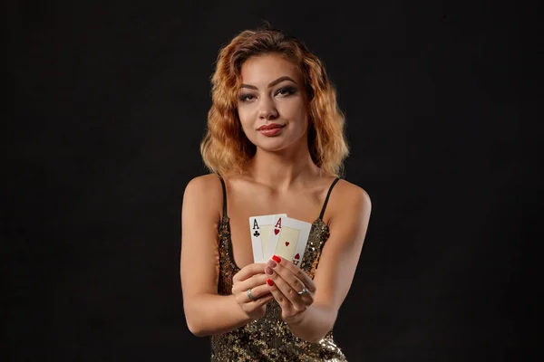 Ginger dívka na sobě lesklé šaty je pózování drží dvě hrací karty v rukou stojí na černém pozadí studia. Kasino, poker. Detailní záběr. — Stock fotografie