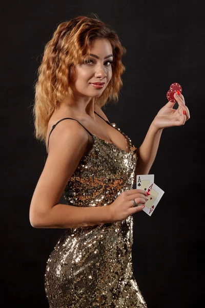 Zázvorová dívka v lesklých šatech pózuje drží dvě hrací karty a žetony v rukou stojí na černém pozadí studia. Kasino, poker. Detailní záběr. — Stock fotografie