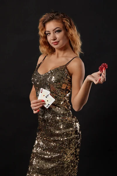 Zázvorová dívka v lesklých šatech pózuje drží dvě hrací karty a žetony v rukou stojí na černém pozadí studia. Kasino, poker. Detailní záběr. — Stock fotografie