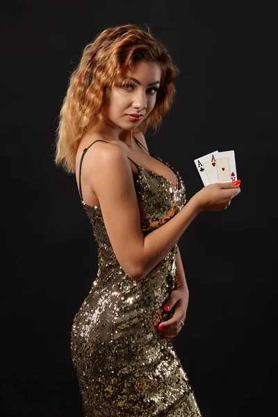 Ginger dívka na sobě lesklé šaty je pózování drží dvě hrací karty v rukou stojí na černém pozadí studia. Kasino, poker. Detailní záběr. — Stock fotografie