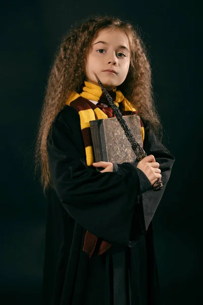 Retrato de una pequeña bruja de pelo largo y castaño vestida con abrigo oscuro, sosteniendo varita mágica en la mano, posando sobre fondo negro del estudio . — Foto de Stock