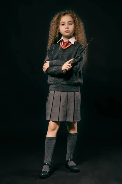 Retrato de una pequeña bruja con el pelo largo y castaño vestido con ropa oscura, sosteniendo varita mágica en la mano, posando sobre fondo negro del estudio . —  Fotos de Stock