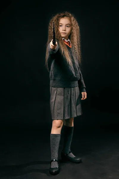 Retrato de una pequeña bruja con el pelo largo y castaño vestido con ropa oscura, sosteniendo varita mágica en la mano, posando sobre fondo negro del estudio . —  Fotos de Stock