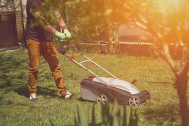 Young worker in casual clothes and gloves is mowing green grass with lawn mower on a yard. Gardening care equipment and services. Sunny day clipart