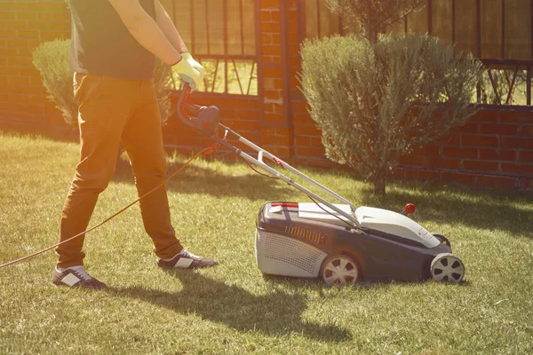 Mann in Freizeitkleidung und Handschuhen mäht Gras mit modernem Rasenmäher auf seinem Hinterhof. Geräte und Dienstleistungen für die Gartenpflege. Sonniger Tag — Stockfoto
