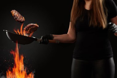 Elleri dövmeli, tayt ve tişört giymiş bir aşçı. Tavayı ateşin üstünde tutmak ve siyah arka planda iki bifteği kızartmak. Yan görünüm