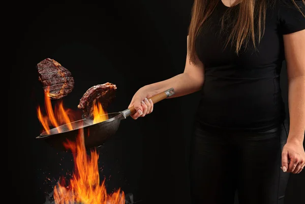 Cuire avec les mains tatouées, vêtu de leggings et t-shirt. Tenir la casserole de wok au-dessus du feu et faire frire deux steaks de bœuf sur fond noir. Vue latérale — Photo