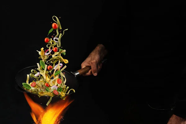 Un homme méconnaissable tenant une casserole de wok au-dessus du feu et cuisinant des pâtes avec des tomates cerises, de l'oignon et du basilic sur fond noir. Gros plan — Photo