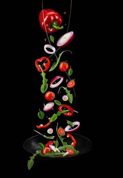 Wok pan, falling ripe tomatoes, red bell pepper, onion, green basil and arugula against black studio background. Poured by a water stream. Close up