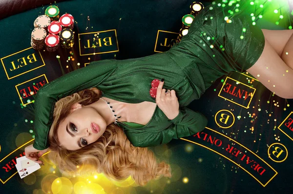 Blonde female in green dress is showing red chips and aces. Lying on playing table with colorful sparkles above her. Poker, casino. Close-up, top view — Stock Photo, Image