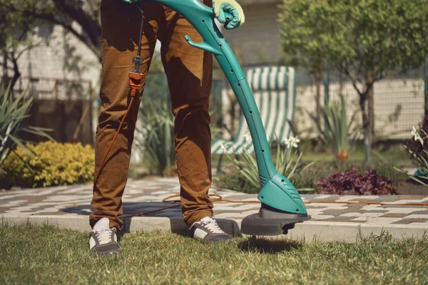 Kerl in lässiger Kleidung und bunten Handschuhen mäht mit professionellem elektrischen Mini-Rasenmäher im Garten seines Landhauses grünes Gras. Nahaufnahme — Stockfoto