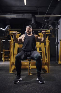 Dövmeli, sakallı, siyah eşofman altı, yelek ve şapka giyen güçlü bir adam. Göğüs presi yapıyor, karanlık spor salonundaki egzersiz makinesinde oturuyor. Tam uzunluk