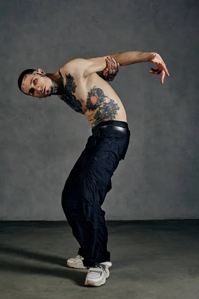 Atletisch getatoeëerde man met naakte romp, baard. Gekleed in zwarte broek en witte sneakers. Dansen tegen een grijze achtergrond. Danszaal, hiphop — Stockfoto