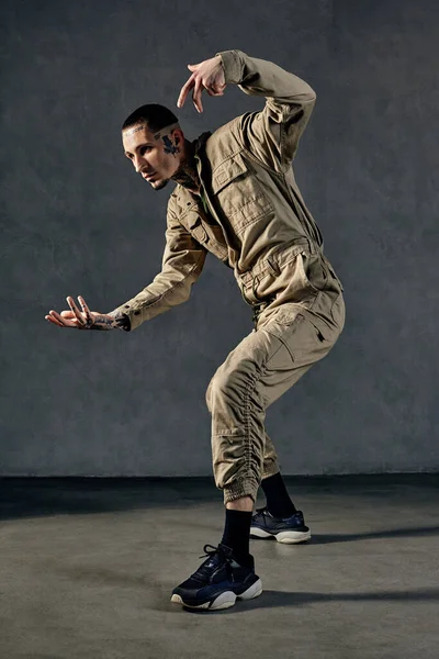 Flexibele performer met getatoeëerd lichaam, oorbellen, baard. Gekleed in kaki overalls en zwarte sneakers. Dansen op grijze achtergrond. Danszaal, hiphop — Stockfoto