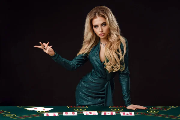 Donna in abito verde sta mostrando qualcosa, appoggiata sul tavolo da gioco con le carte su di esso, in posa su sfondo nero. Poker, casinò. Primo piano, copia spazio — Foto Stock