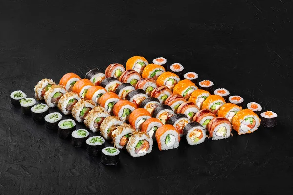 Somon, avokado, salatalık ve diğer malzemelerin yer aldığı çeşitli suşi dürümleri ve maki üçgenleri siyah arka planda sunulmaktadır.