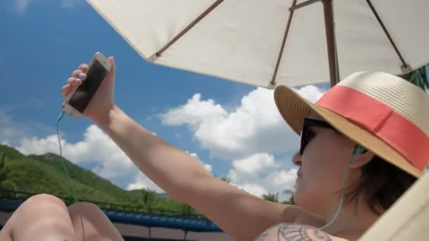 Una mujer con sombrero bajo sombrilla en una tumbona toma fotos de sí misma con un teléfono celular — Vídeos de Stock