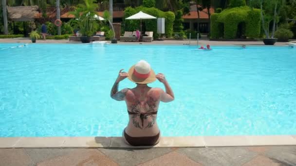 A back view of a tattooed woman in a hat is sitting by the swimming pool and looking around — Stock Video