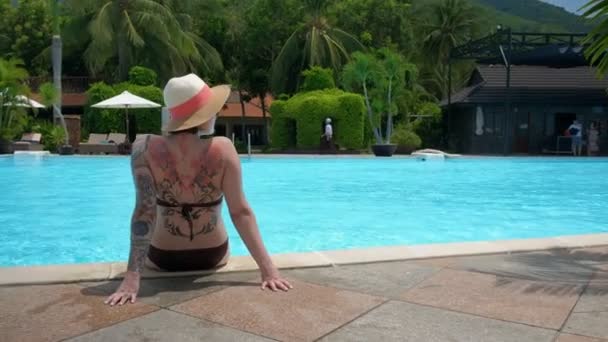 Uma visão traseira de uma mulher tatuada em um chapéu está sentada à beira da piscina e olhando ao redor — Vídeo de Stock