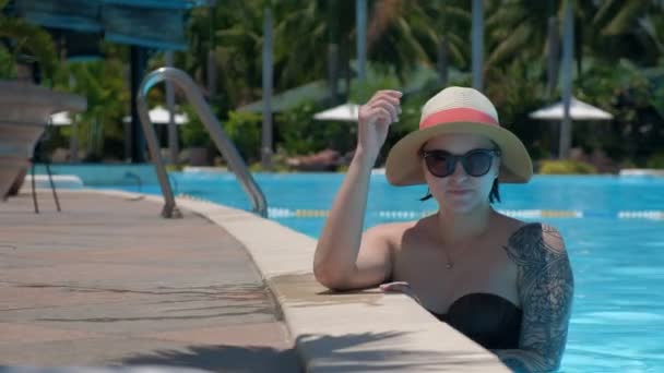 Hermosa joven con gafas de pie en la piscina mira a la cámara y corrige sombrero de mano — Vídeo de stock