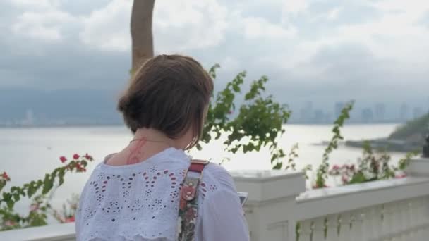 Μια πίσω προβολή ενός τατουάζ κορίτσι περπάτημα και πληκτρολόγηση σε smartphone της με έναν ωκεανό, αστικό τοπίο και τα βουνά στο φόντο — Αρχείο Βίντεο