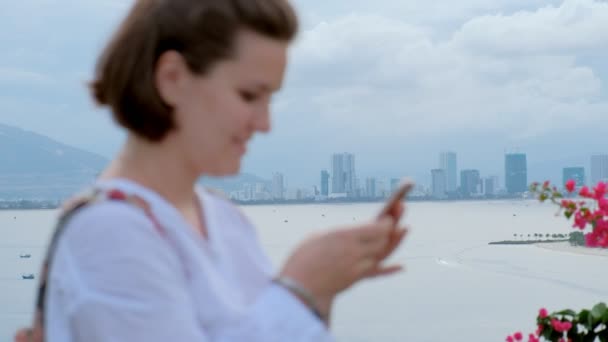 Un primer plano desenfocado de una hermosa mujer joven escribiendo en su teléfono inteligente con un océano y el paisaje urbano en el fondo — Vídeo de stock