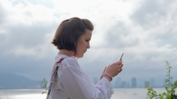 Un primer plano lateral de una hermosa joven feliz charlando en su teléfono inteligente con un cielo azul y el océano en el fondo — Vídeos de Stock