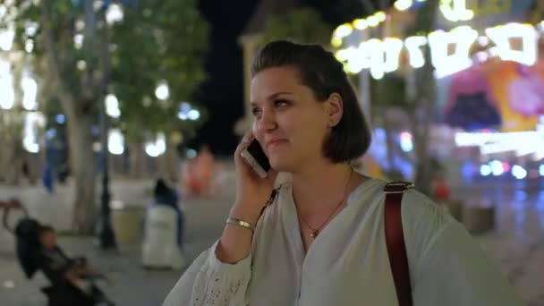 A steadicam lövés egy csinos, fiatal nő beszél a telefonon, és mosolyogva, éjszaka, a háttérben egy izzó vidámpark — Stock videók