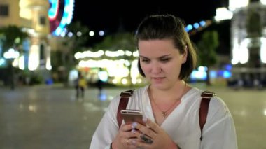 Güzel ve genç birbayan manifatura onun akıllı sonra kameraya bakıyor ve arka plan üzerinde parlayan bir eğlence parkı ile gülümseyen fotoğrafını yakın çekim