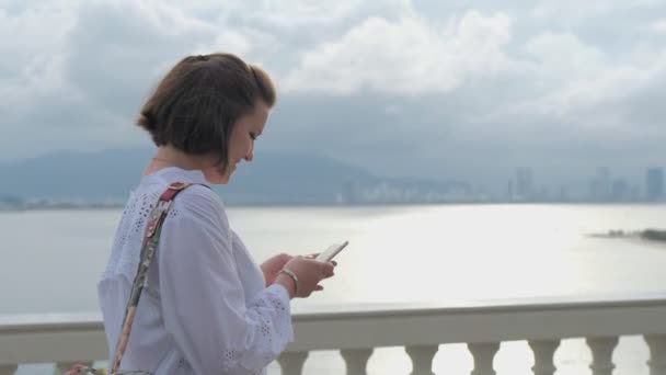 Un primer plano de una hermosa mujer joven caminando y escribiendo en su teléfono inteligente y luego mirando alrededor del océano y paisaje urbano en el fondo — Vídeos de Stock