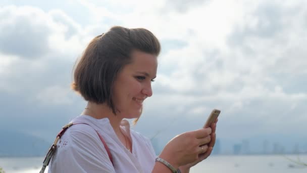 Uma foto de close-up de uma bela jovem digitando em seu smartphone com um oceano e paisagem urbana no fundo — Vídeo de Stock