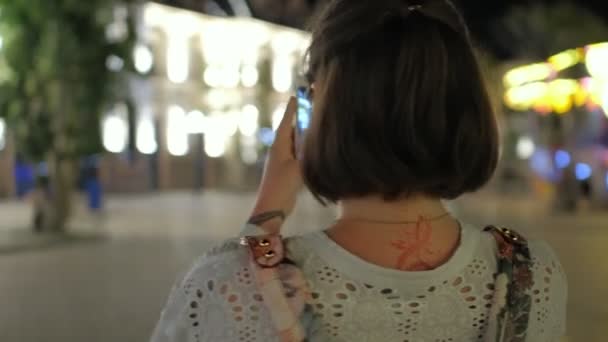 Una foto steadicam de una hermosa joven sonriente tomando fotos con su teléfono inteligente en un parque de atracciones por la noche — Vídeos de Stock