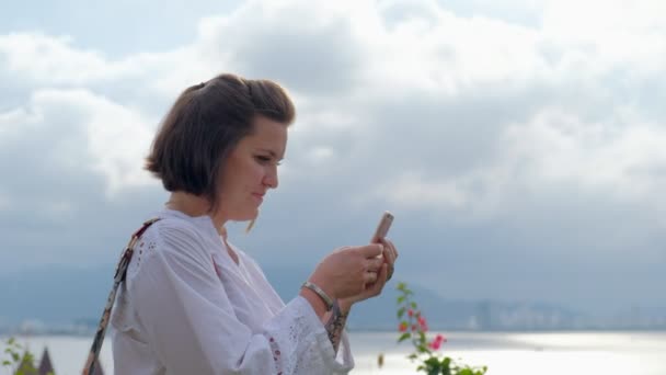 Un primer plano lateral de una joven muy feliz charlando en su teléfono inteligente con un cielo azul y el océano en el fondo — Vídeo de stock